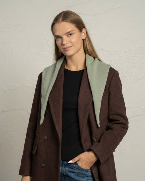 A woman wearing a dark brown coat, black top, and jeans poses with a light green knitted mini bandana draped over her shoulders. She stands against a textured light-colored wall, looking toward the camera with a slight smile.