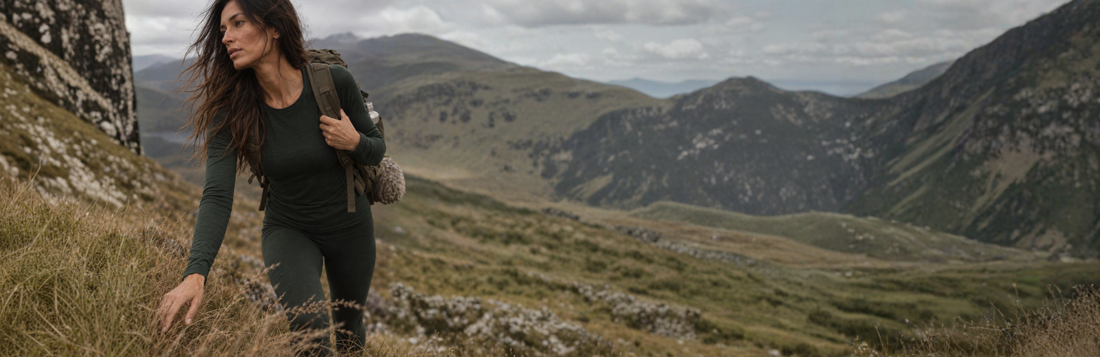 A woman with long dark hair hikes uphill near a large, textured boulder, wearing a dark green long-sleeved top and matching leggings. She carries a large olive-green backpack and looks out across a sweeping landscape of rolling hills beneath a partly cloudy sky.