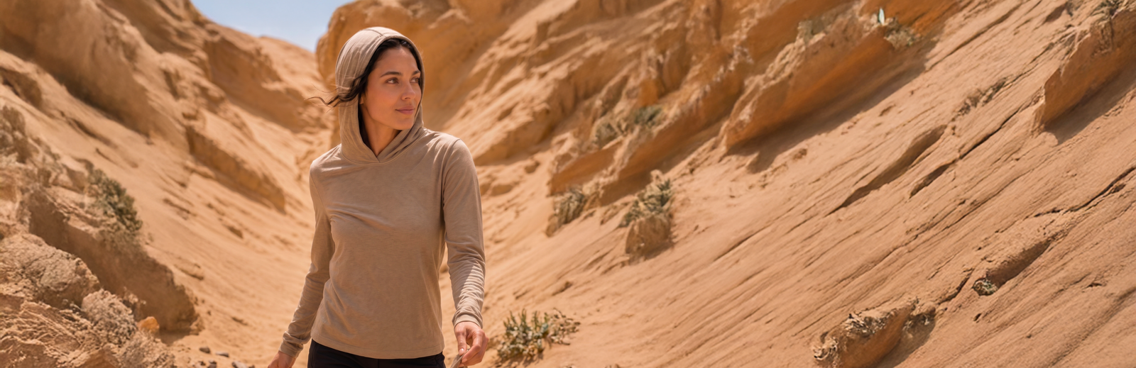 A woman wearing a lightweight, beige long-sleeve sun hoodie walking through a narrow, sandy desert canyon.