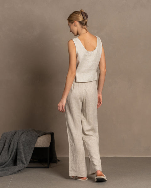A woman with blonde hair in an updo wears a natural linen sleeveless crop top and matching pants, standing with her back to the viewer. A gray blanket is draped over a small bench in the lower left corner, against a muted brown textured background. 