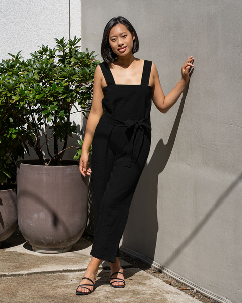 A person with short, dark hair and what appears to be Asian features is leaning against a textured wall. They are wearing a black jumpsuit with wide straps and a tied waist, paired with black strappy sandals. Two large potted green plants are visible to the left.