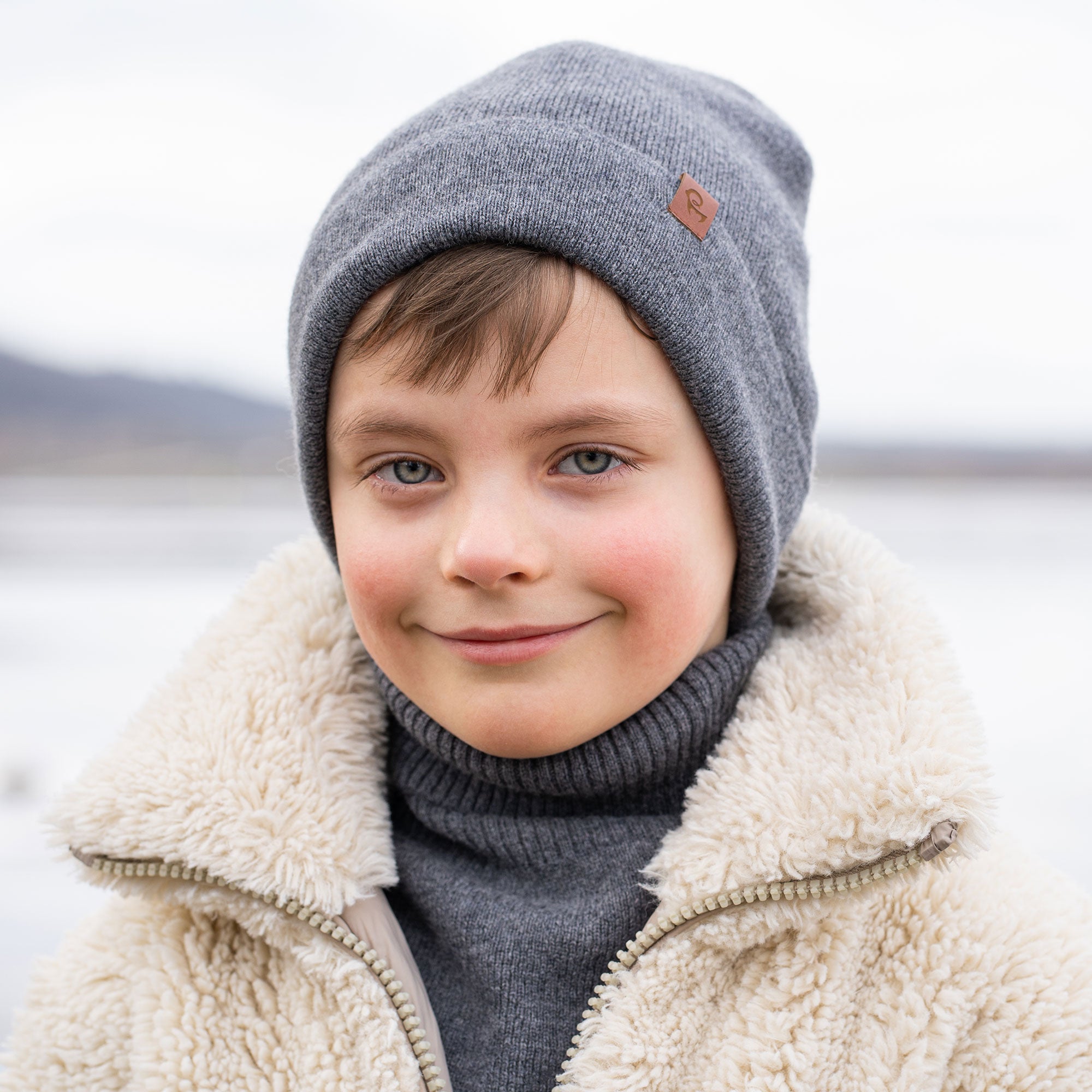 A young child with short brown hair and a wide smile, is dressed in winter clothing. They are wearing a beige beanie, a beige turtleneck sweater, and a cream-colored sherpa jacket, with beige knitted gloves. A snowy, blurred background with what appears to be a body of water is visible behind them.