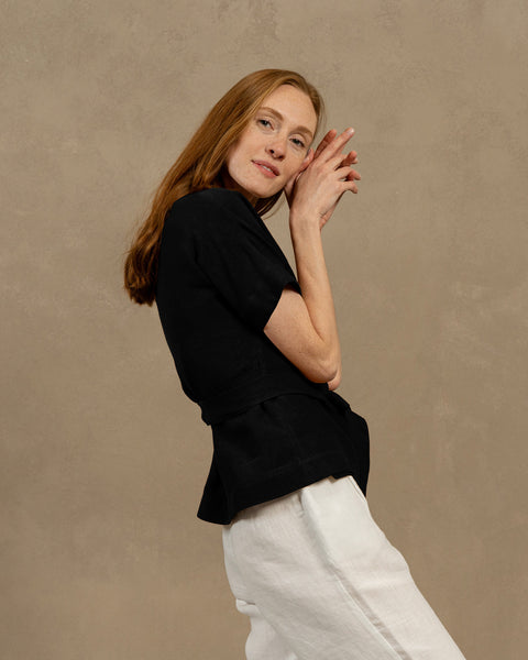 A fair-skinned person with long, auburn hair, wearing menique black V-neck linen wrap top with short, wide sleeves and light-colored pants. They are holding the ends of the top&#39;s tie in front of them, forming a bow.