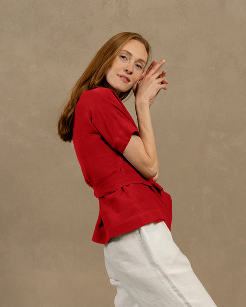 A fair-skinned person with long, auburn hair, wearing pure red V-neck linen wrap top with short, wide sleeves and light-colored pants. They are holding the ends of the top&#39;s tie in front of them, forming a bow.