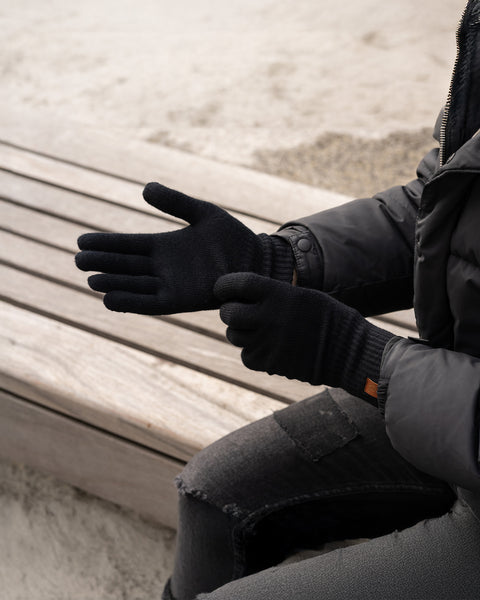 A person wearing black knit gloves from menique, while sitting on a wooden bench outdoors. The gloves appear to be snug-fitting and warm.