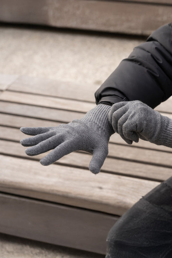 A person wearing gray ribbed knit merino wool gloves, sitting on a slatted wooden surface. The gloves have a snug fit and appear to be warm. A small brown tag is visible on the wrist of the right glove.