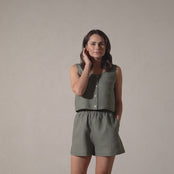 A woman with shoulder-length brown hair models a sleeveless, button-up stone green top and matching linen shorts, slowly turning in a full circle against a plain background in a short video.