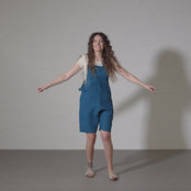 A woman with long, curly brown hair dances energetically in a short video, wearing a white t-shirt underneath short linen romper. She points with both hands and moves against a plain studio backdrop.