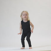 A young child with blonde hair, wearing a black menique top and black pants, stands against a white background with their arms raised above their head. The child is looking directly at the camera.