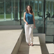 A woman with shoulder-length brown hair models a menique linen one-shoulder top and white pants, walking down a short set of concrete steps outdoors in a short video.
