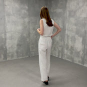 This is a short video of a fair-skinned woman with long reddish-brown hair modeling a pure white linen sleeveless vest and matching wide-leg pants. She is wearing black open-toe heeled sandals and poses in a three-quarter view, looking towards the camera with a slight smile, her right hand casually placed in her pocket. The setting is a simple studio with textured gray walls and floor.