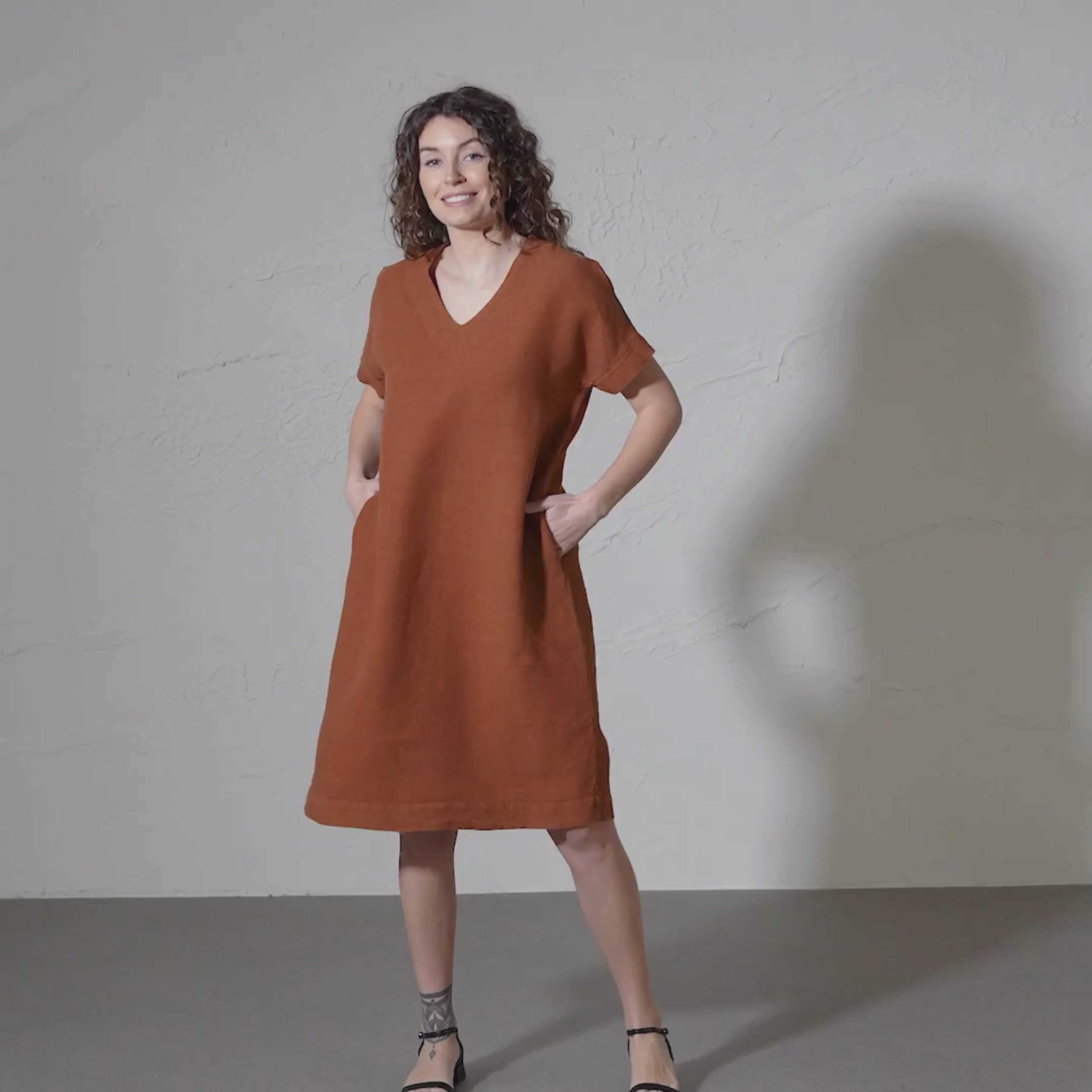 A woman with curly brown hair models a loose-fitting, short-sleeved, almond brown colored dress with a V-neck and side pockets against a textured light gray wall.