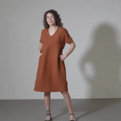 A woman with curly brown hair models a loose-fitting, short-sleeved, almond brown colored dress with a V-neck and side pockets against a textured light gray wall.