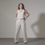 A smiling woman with long, curly brown hair models a linen sleeveless top and matching menique linen wide-legged pants, posing with her hands on her hips in a short video against a textured off-white wall.