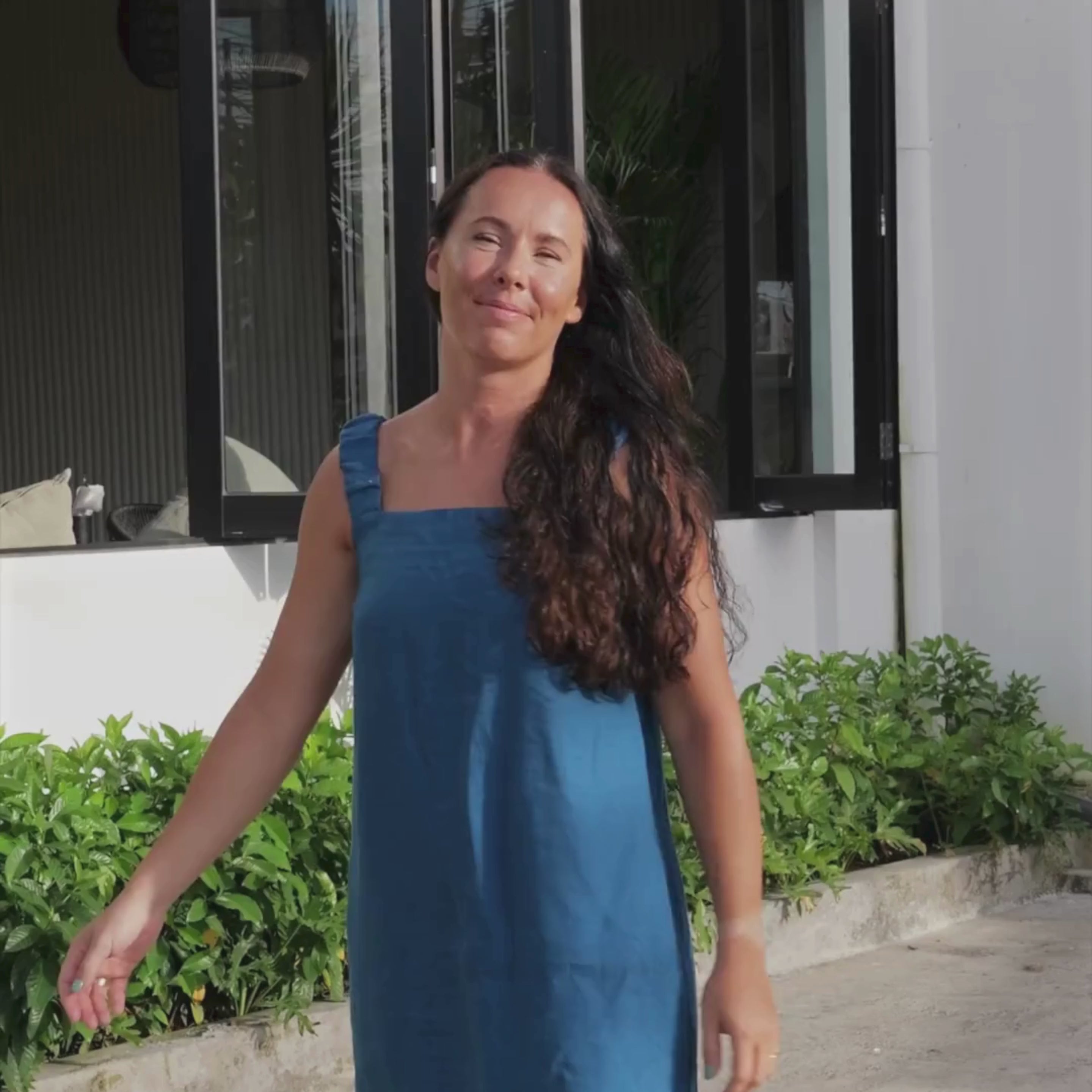 A woman with long, dark wavy hair and light to medium skin tone smiles and walks towards the camera in a short video clip. She is wearing a sleeveless, cobalt blue linen dress with wide straps. She appears to be outdoors, with green foliage visible in the foreground and a building with dark-framed windows in the background.
