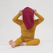 A child with fair hair and blue eyes, wearing a royal cherry hood and a yellow long-sleeved top, looks at the camera with a slight smile. The child's left hand is resting against their cheek. The background is white. 