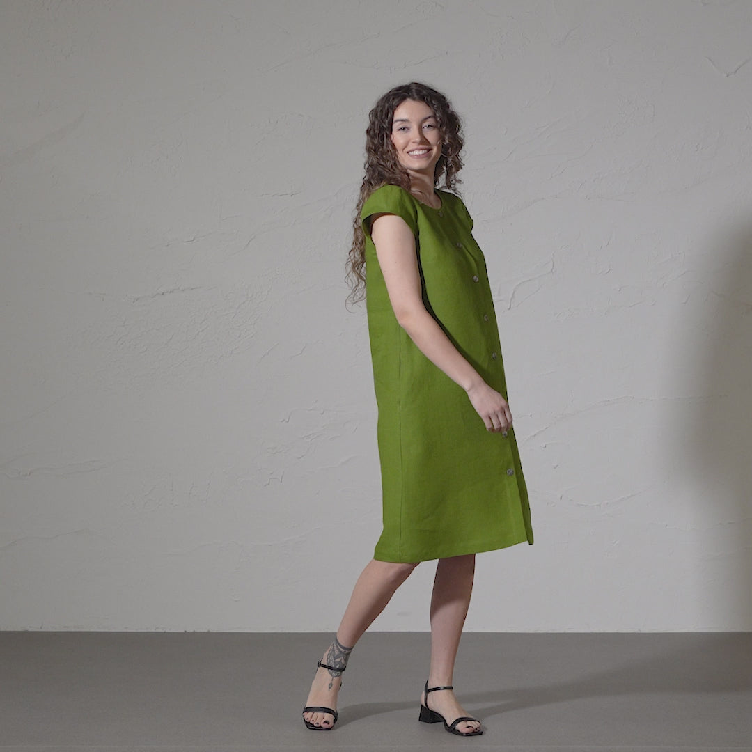 A woman with curly brown hair smiles and models a short-sleeved, forest green midi linen dress with a button-down front against a textured gray backdrop in a short video. 