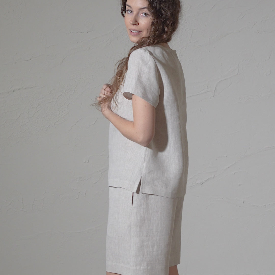 A woman with long, curly brown hair models a natural short-sleeved linen top and matching pants against a textured gray backdrop in a short video. They glance over their shoulder and smile.