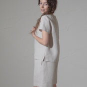 A woman with long, curly brown hair models a natural short-sleeved linen top and matching pants against a textured gray backdrop in a short video. They glance over their shoulder and smile.