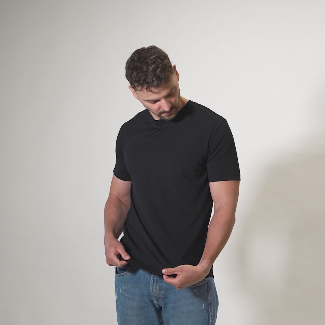 A man with short brown hair and a beard, stands and later sits on a black stool. He is wearing a black, then dark green and blue t-shirt and jeans, looking at the camera with his hands clasped. The background is a solid white.