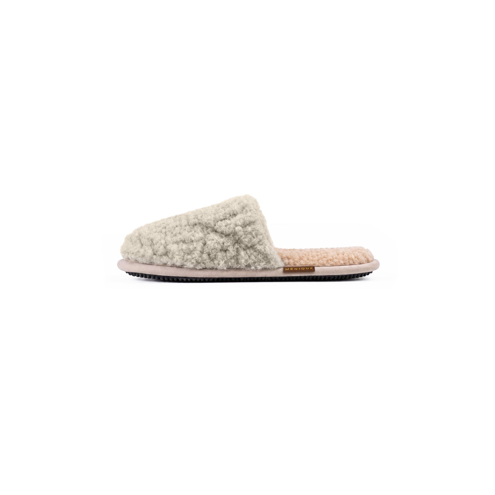 A person sitting on a bed wearing beige, fuzzy slip-on slippers with a small brown "MENIQUE" tag, resting their feet on a textured rug.