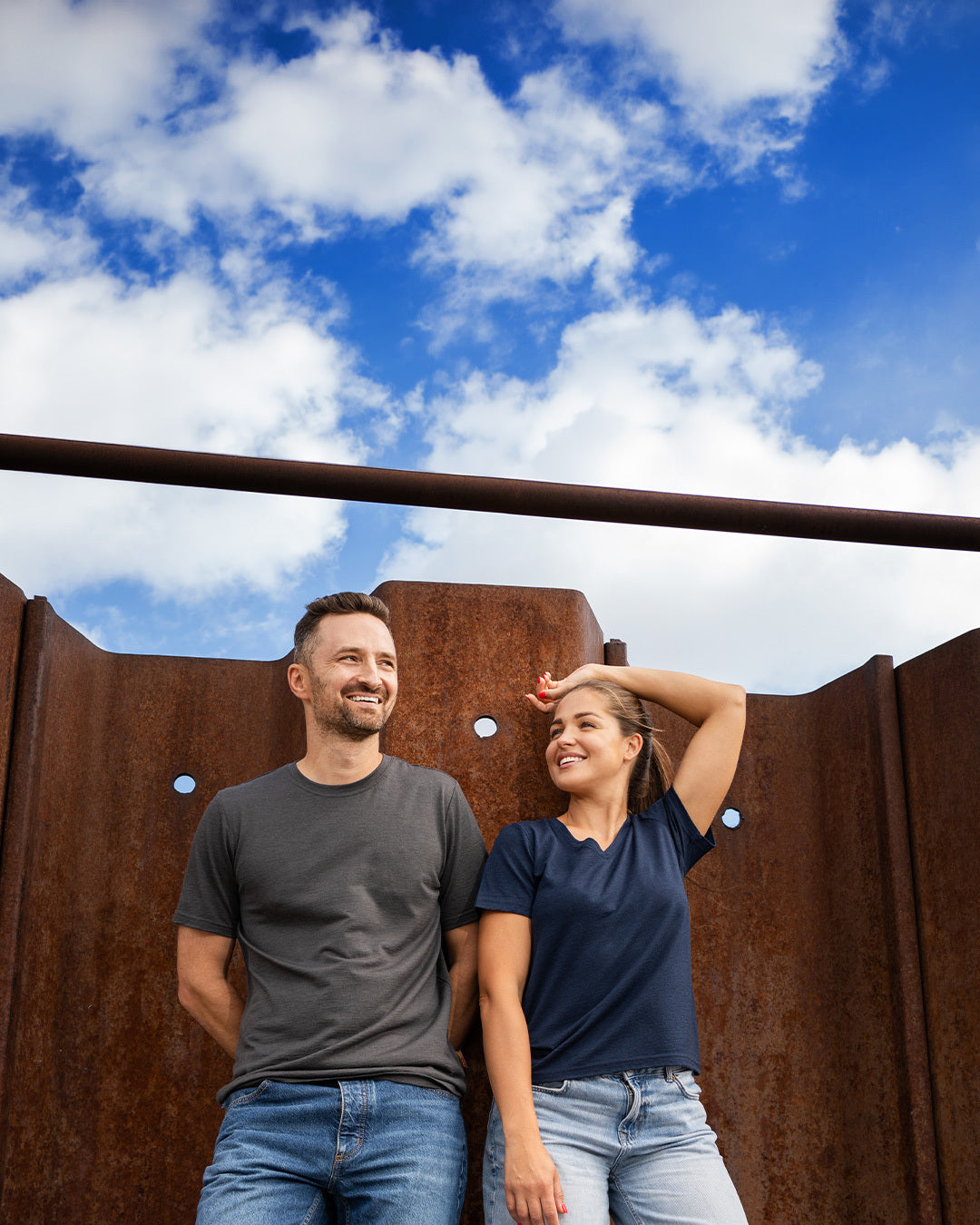 A wide shot of a smiling man and woman standing together outdoors in front of a tall, rusty corrugated metal wall under a partly cloudy sky. The man, on the left, is looking off-camera and wearing a charcoal grey short-sleeve crew neck t-shirt and blue jeans, with his hands behind his back. The woman, on the right, is looking at him with a smile and is wearing a navy blue short-sleeve v-neck t-shirt and light blue jeans, with her left arm resting on the wall above her head.