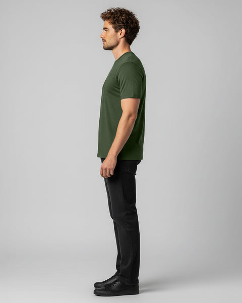 A side view of a man wearing a green T-shirt and black jeans, standing against a neutral gray background.
