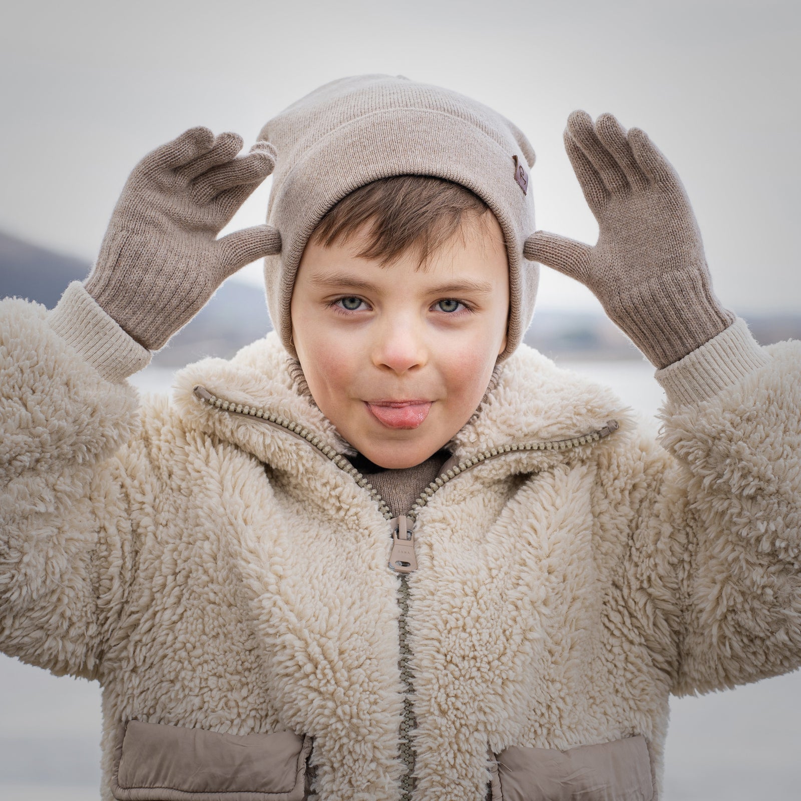 Child wearing beige sherpa jacket, knit hat and gloves, playfully posing outdoors in winter