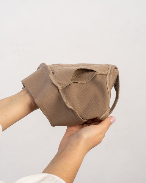 Hands holding a light brown bralette, possibly made of soft, lightweight fabric, against a plain light background.