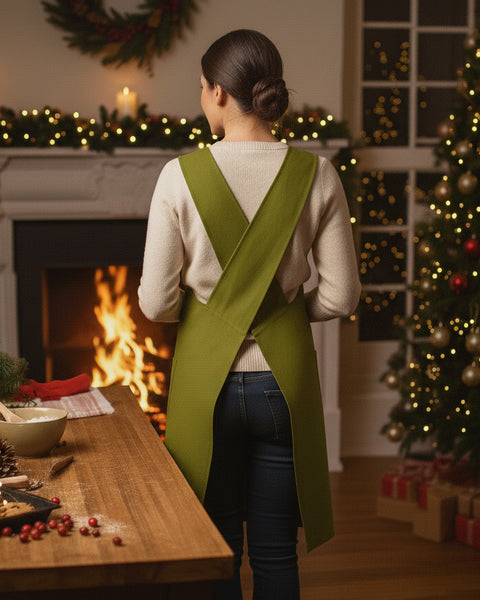 Woman wearing a green linen cross-back apron, standing in a cozy Christmas kitchen near a fireplace and decorated tree with holiday lights.