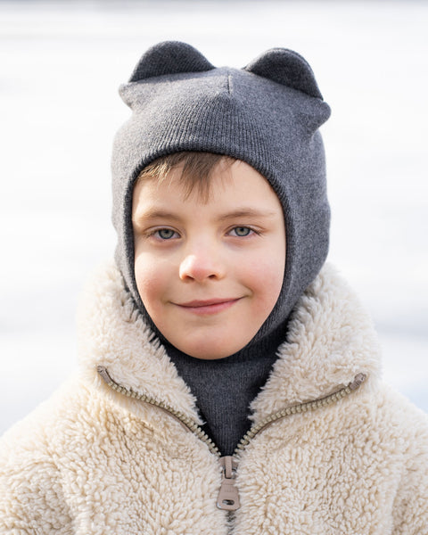 A child with light brown hair and blue eyes wears a dark gray knit balaclava with two small, rounded ear-like protrusions on top. They also wear a light-colored, fuzzy or shearling-like jacket with a zipper partially open, revealing a dark gray turtleneck underneath. The child is looking directly at the camera with a slight smile. The background is blurred, showing a light, possibly icy or snowy, outdoor environment.