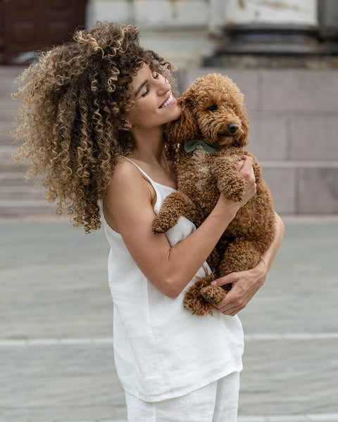 Beautiful woman posing in the city, wearing pure white linen slip top and pure white linen pants with black sandals, holding cute brown poodle in her hands.