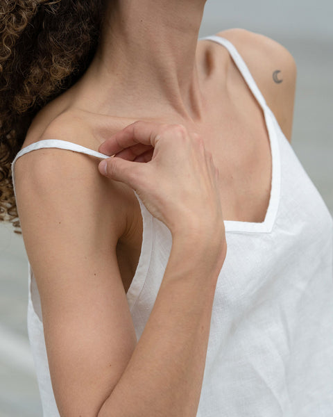 Close-up of a woman from front, wearing a pure white linen slip top, on her left shoulder minimalist tattoo of a moon.