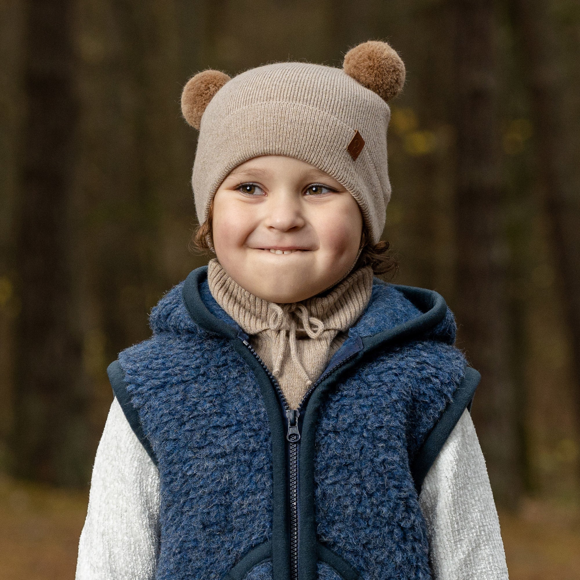 A young child with blue eyes and a wide smile, is looking directly at the viewer while peeking over a metal playground railing. They are wearing a beige knit hat with brown pom-pom ears, beige mittens, and a light pink sherpa-style coat.