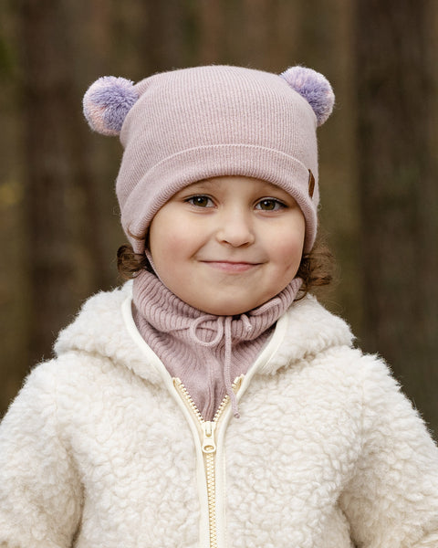 A young child with light brown curly hair and a subtle smile, is looking directly at the viewer. They are wearing a light pink knit hat with two lighter pink pom-pom ears, a matching pink turtleneck or neck warmer, and a cream-colored sherpa jacket with a zipper. The background is a blurred forest.