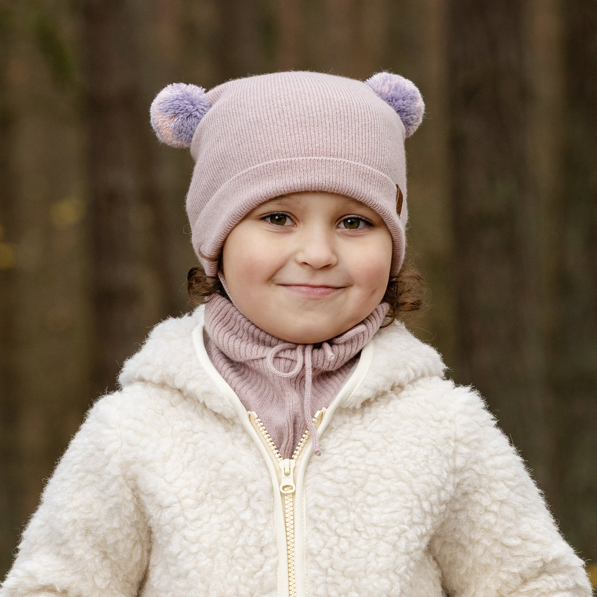 A young child with blue eyes and a slight smile, looks directly at the viewer. They are wearing a beige knit hat with brown pom-pom ears, a beige turtleneck or neck warmer, and a light pink sherpa-style coat. The background is a blurred outdoor scene with muted green and brown tones.