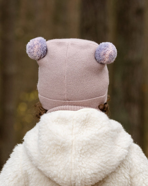 The back of a child&#39;s head and shoulders are shown, with the child wearing a light pink knit hat adorned with two pink and purple speckled pom-poms on top. They also wear a lighter pink turtleneck, and the hood of a cream-colored sherpa jacket is visible, set against a blurred forest background.