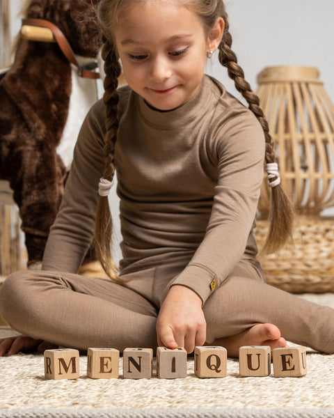 A girl, playing on the carpet with wooden cubes, wearing the menique Kids&#39; Merino 160 Pants Beige color.