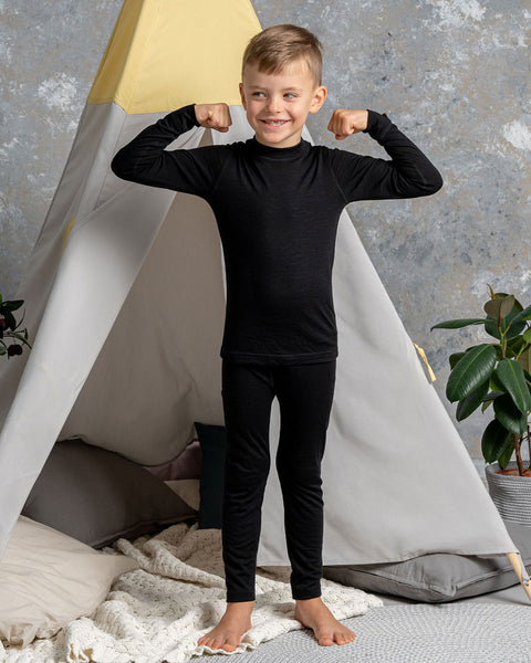 a boy wearing the menique Kids&#39; Long Sleeve Set 160 Merino in Black color. He is standing with his arms raised ant bent, acting strong.