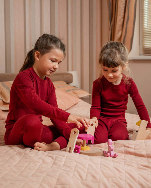 Two young children are kneeling on a light pink quilted bed, playing with wooden toys. Both children are wearing matching long-sleeved, deep red pajama sets. The child on the left, with dark hair tied back in a ponytail, is looking down at the toys and appears to be interacting with a small pink table and chairs.