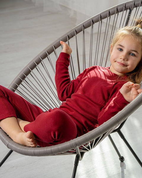 A young child with light hair pulled back into a ponytail is sitting comfortably in a modern, gray string-style chair. The child is wearing a long-sleeved royal cherry top and matching red pants, and is looking towards the viewer with a slight smile. 